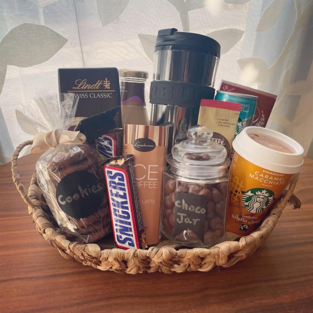 Woven gift basket filled with coffee drinks, chocolates, and a black travel mug.