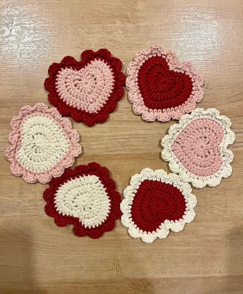 Six heart-shaped crochet coasters in red, pink, and cream on a wooden table.