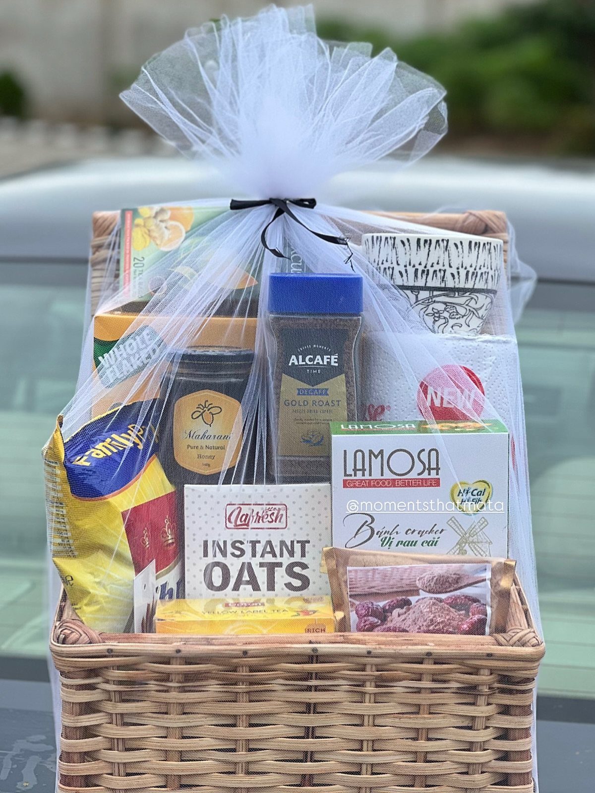 Wicker gift basket filled with food items wrapped in clear cellophane and tulle.