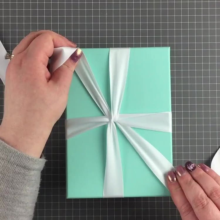 Hands tying a white satin ribbon around a turquoise gift box on a grid mat