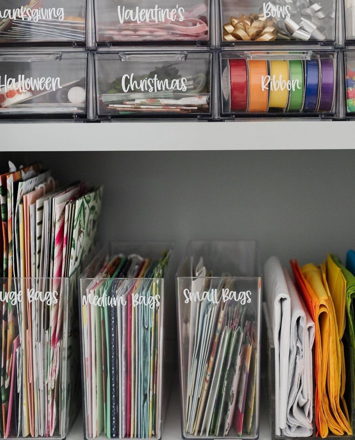 Clear plastic storage bins organizing colorful wrapping paper, gift bags, and ribbons on shelves.