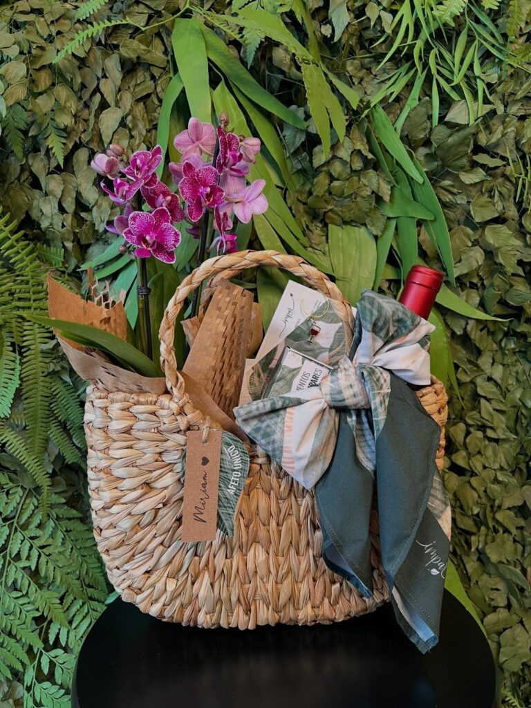 Woven gift basket with wine, purple orchids, and a blue patterned bow.